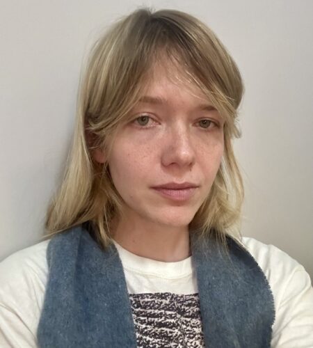 Photo of the author in a white Tee shirt and blue scarf, standing in front of a neutral wall.