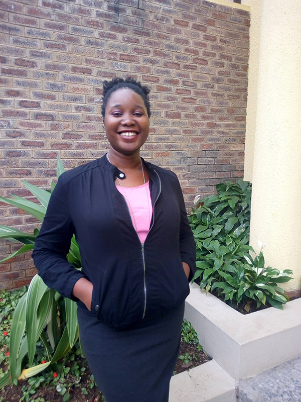 Akumbu Uche, a Black woman in black skirt and jacket over a pink tee, smiles in front of some green plants and a brick wall. Her hair is pulled into a ponytail and her hands are in her pockets.