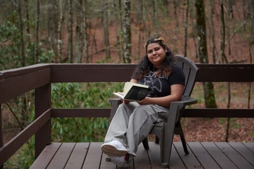 Shlagha Borah, a woman raised in India, sits on a fenced deck in a brown deck chair, with trees behind her. She is holding a book open and wearing loose baggy pants and a black T-shirt with a white stencil.