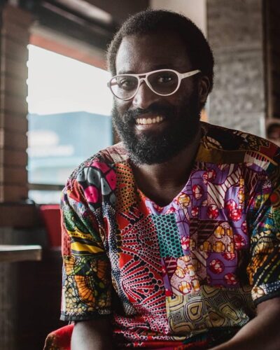 Mugabi Byenkya, a Nigerian man with a full beard, white-rimmed glasses, and a colorful shirt, smiles with a brick wall and window behind him.