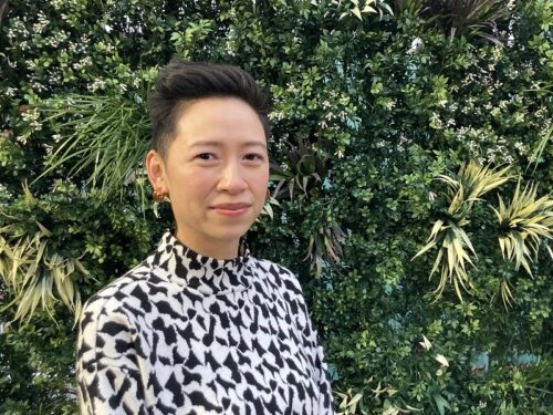 Lisa Low, an Asian American woman with short hair, wearing a black and white decorative shirt, stands in front of greenery