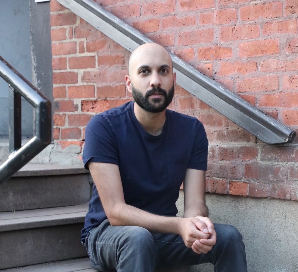 Ravi Mangla, an Asian-American man with shaved head and short black beard, sits on some outdoor steps in front of an industrial brick wall.