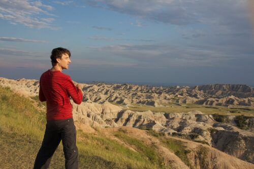 A white man in a red long-sleeve shirt and dark pants looks out over a vista of craggy rocks and grass.