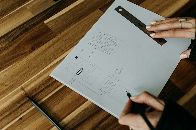 A woman's hands hover over a piece of paper with a pencil and ruler. On the paper are pencil sketches of wireframes for what looks like an app or part of a website. The paper sits on a wooden surface.