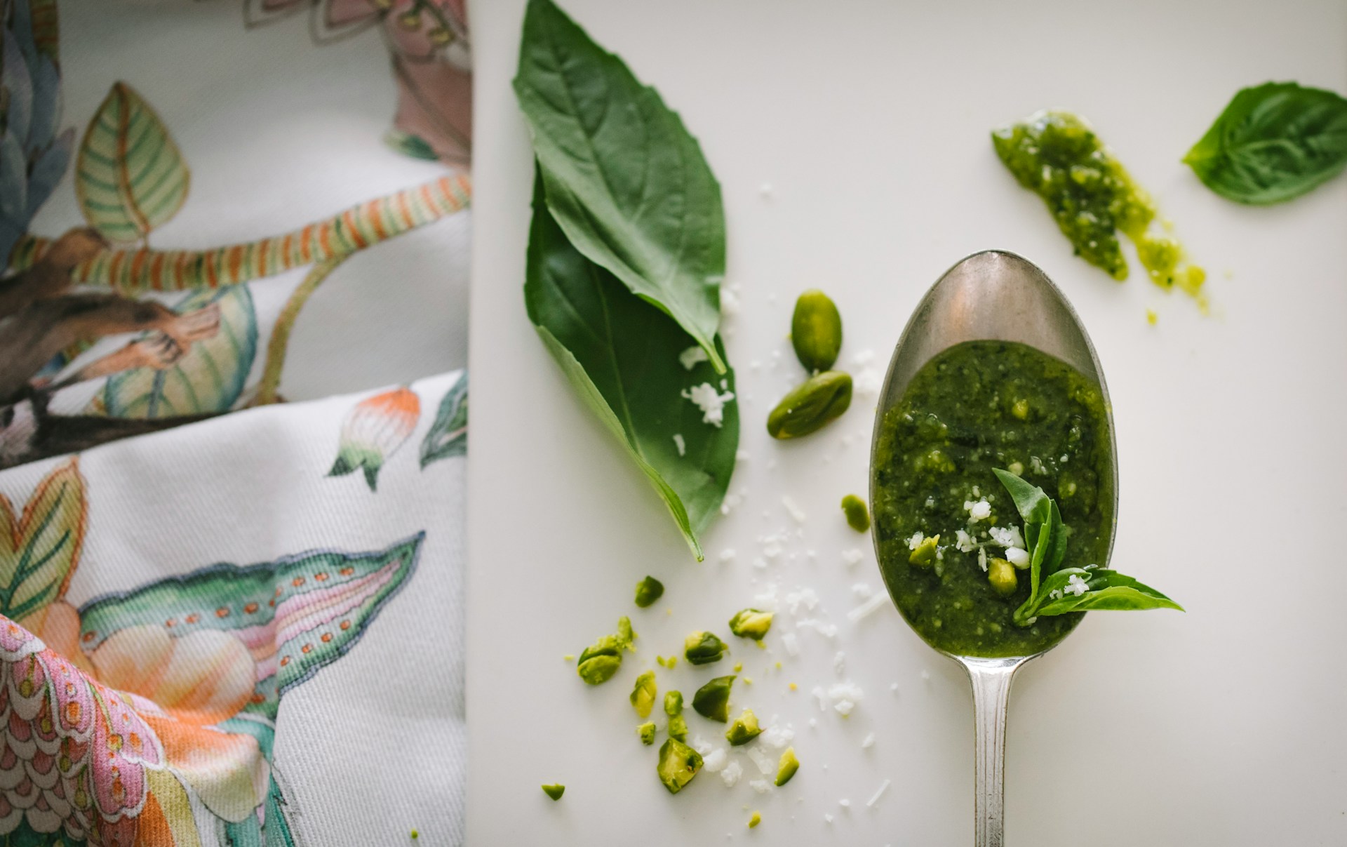 Flatlay of pesto ingredients: basil leaves, pistachios, cheese, and a spoonful of pesto against a fabric background.