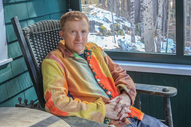 David DeGusta, a white man with red hair and blue eyes wearing a multicolored orange and yellow sweater, sits on a rocking chair on a screened porch, with trees and snow outside.