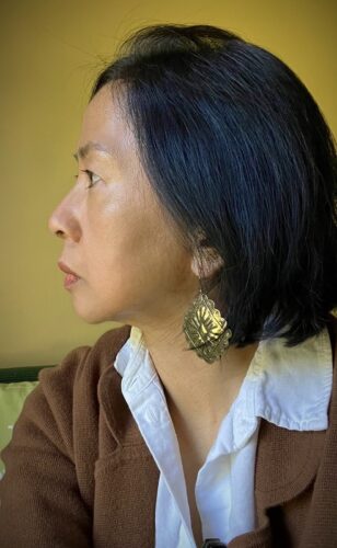 An Asian American woman in profile, looking toward our left. She is wearing a large diamond-shaped gold earring and a white button-down shirt and brown sweater. The wall behind her is chartreuse colored.