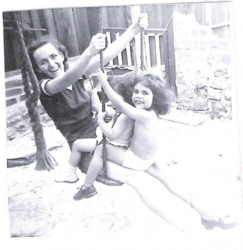 A black and white photo of a woman and two girls on a porch swing. The girls, shirtless, sit on their mother's lap, facing her, and one looks at the camera and smiles.