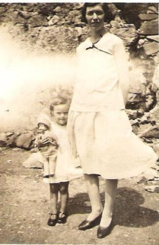 A sepia-toned old picture of a woman and a young girl holding a doll, in front of a rough rock wall.