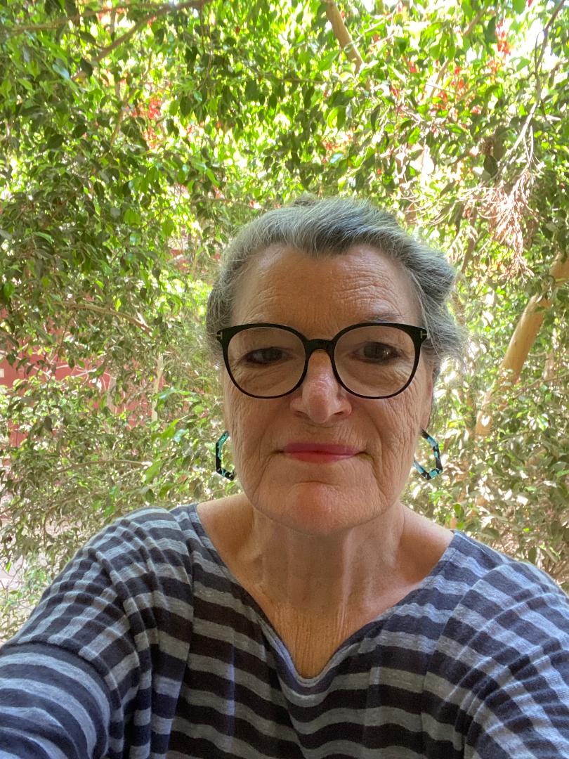 Patricia Newberry, a gray-haired white woman wearing black-rimmed glasses, geometric earrings, and a striped shirt, stands in front of leaves and greenery.