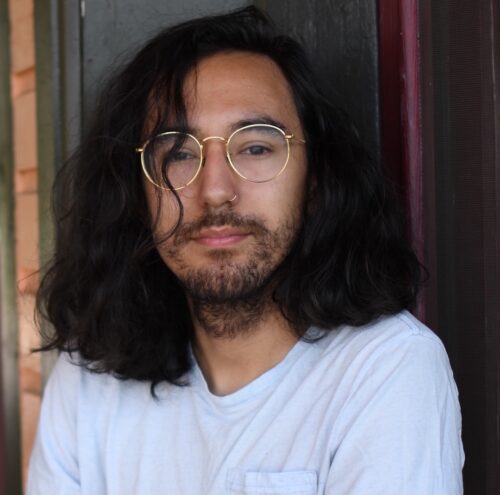 A Latinx nonbinary person with shoulder-length dark hair, gold-rimmed round glasses, a nose ring, a beard, and a white T-shirt.