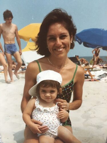 Color photograph of a woman with brown hair in a green and black swimsuit holding a toddler in a white sideways hat and a swimsuit that's white with red dots. Beach umbrellas, beachgoers, and the sea are behind them.