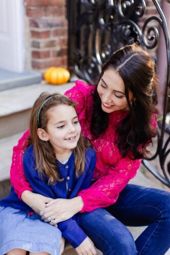 A woman with dark hair, a pink sweater, and jeans, has her arm around a young girl with light brown hair in a headband, a blue button-down sweater, and a blue skirt. They are both smiling, and they sit on a stoop with a wrought-iron fence, small pumpkin, and brick wall behind them.