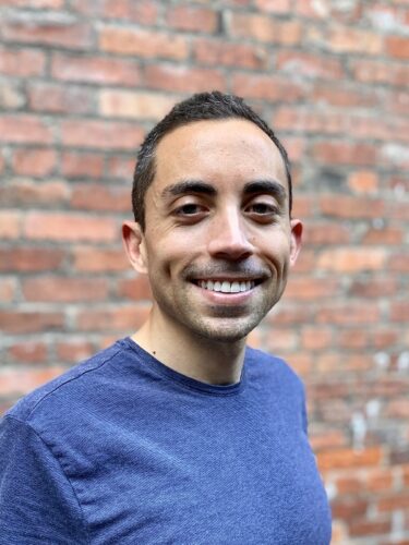 Matthew Torralba Andrews, a multiethnic man wearing a blue T-shirt stands in front of a red brick wall. He has close-cropped dark hair and is smiling.