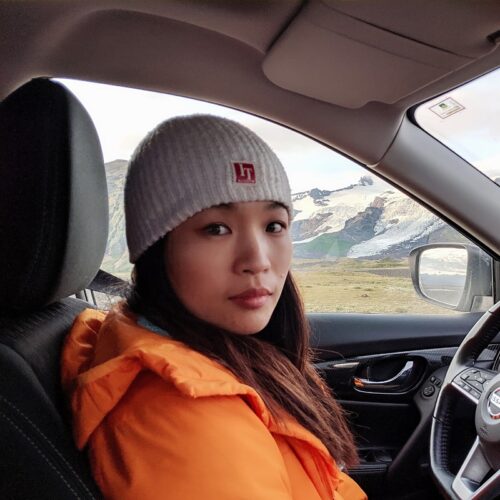 Claire Leng, an Asian American woman, sitting in the driver's seat of a car and wearing an orange jacket and cream-colored knit hat. Mountains and a plain are in the window behind her.