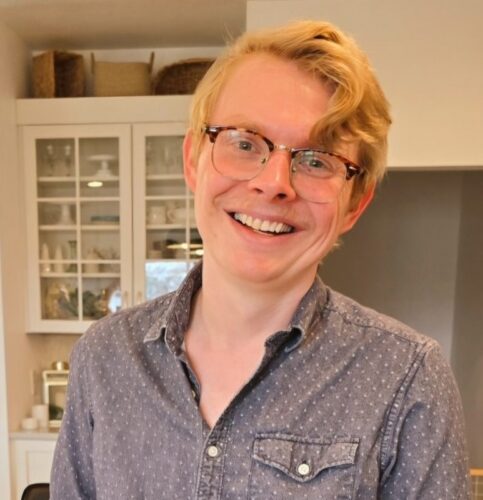 Will Musgrove, a white man with blond hair and glasses horn-rimmed on top. He's wearing a gray button-up collared shirt with white dots and a pocket, and a glass-doored built-in china cabinet and straw baskets are behind him.