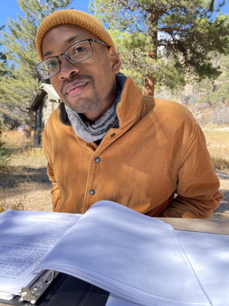 Malik Thompson, a young Black person wearing black glasses, a brown corduroy work jacket, scarf, and matching knitted cap and sitting at a table in a green outdoor space in front of an open book.