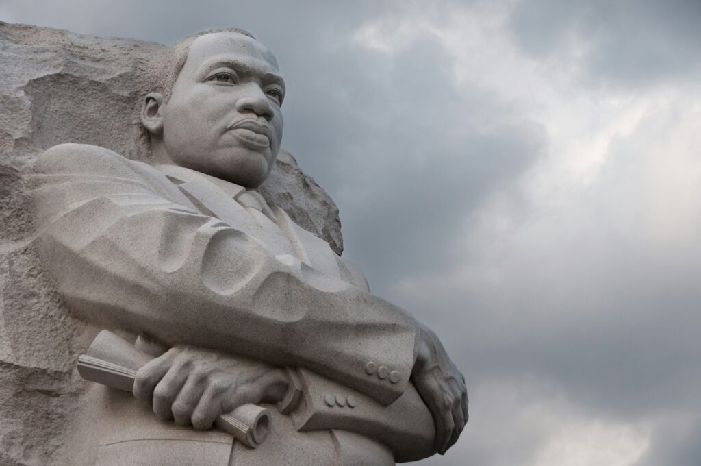 Statue of MLK Jr carved from a stone slab, against a grey, cloudy sky.