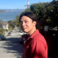 Andy Sia stands in a quiet road with a blurred slope and water behind him. He's wearing a red collared, button-down shirt.