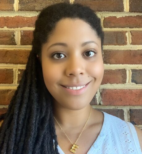 Erica Dawson, a Black woman with long braids over her right shoulder, with a gold necklace that says E and a light-blue shirt. She is smiling and standing in front of a red-brick wall