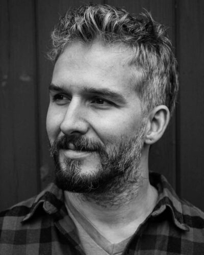 A black and white photo of John Howard, a Colombian American man with a beard, wearing a plaid shirt and smiling slightly as he looks off to the left of the camera.