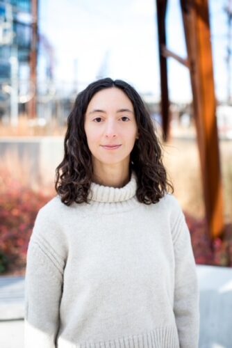 Lily Meyer, a white woman with a white wool turtleneck, stands in front of a blurred winter scene with waters, trees, and shrubs.