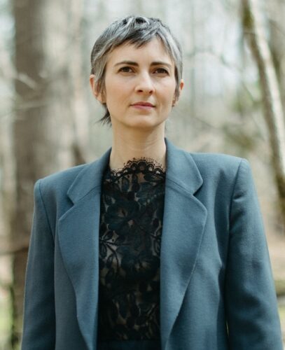 Sarah Rose Nordgren, a white woman with gray and black hair, wearing a blue-gray blazer and lacy black shirt. There are blurred bare trees behind her.