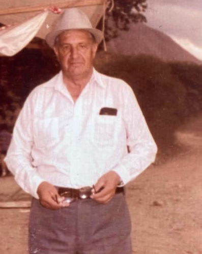 A faded color photo of a Colombian man in a fedora, standing underneath a canvas, with tall bushes and a mountain in the distance.