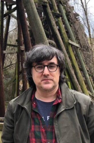 Neil Serven, a white man with dark hair and glasses, is standing outside in front of a wooden structure. He is wearing a dark jacket, plaid shirt, and blue T-shirt.