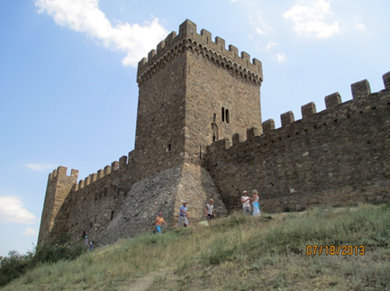 Wall and tower of the fortified city of Sudak. Parapets line the top of the wall. People walk below on a grassy hill.