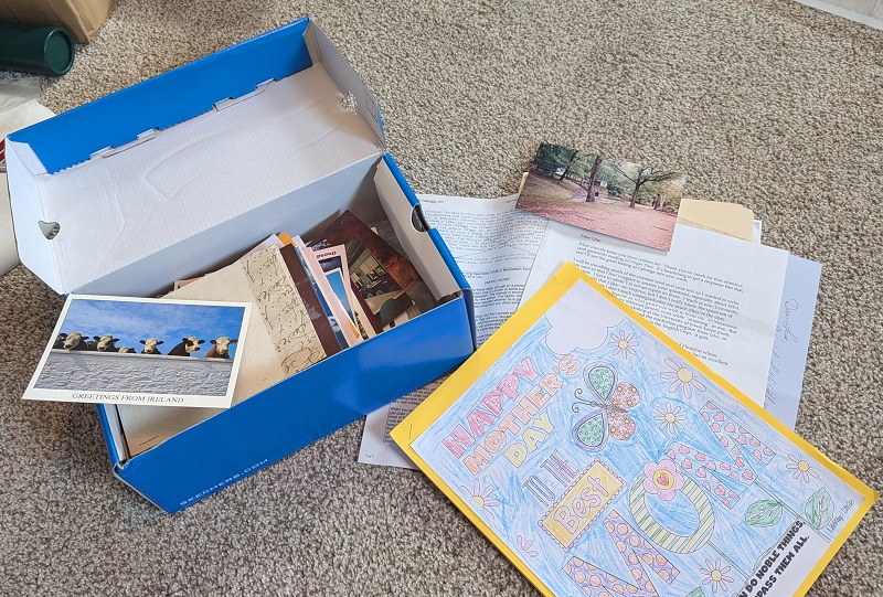 A shoebox with a blue exterior is full of postcards, envelopes, and pictures, and next to it are a few other pieces of paper and a child's Happy Mother's Day coloring.