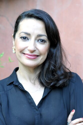 An Iranian American woman smiling with a reddish wall blurred behind her. She is wearing a button-down collared shirt and crossing her arms.