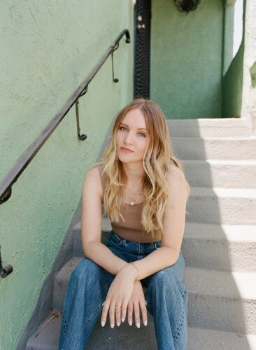 A white woman with long blond hair sitting on outdoor steps with a railing next to her. The wall next to and behind her is a textured light green. She is wearing a beige tank top and jeans, and her hands are crossed with her elbows on her knees.