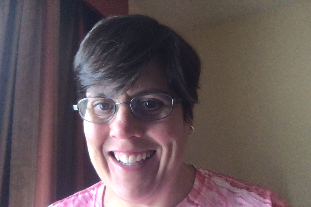 Dinah Cox, a white woman with short brown hair and oval glasses, smiles in front of a plan background. She is wearing a pink t-shirt and small hoop earrings.