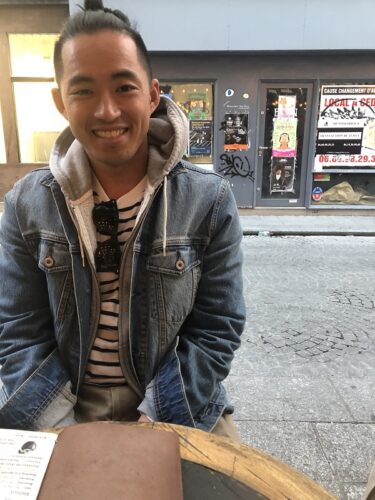 Patrick J. Zhou, an Asian American man, sits at an outdoor table. He's wearing a denim jacket, gray hoodie, and striped white-and-black shirt with sunglasses tucked at the neck. There's a street and storefront behind him.