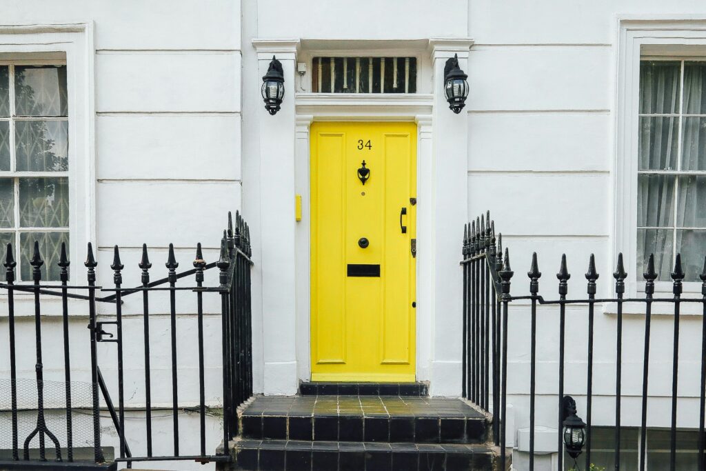 A yellow-painted door on a white London-style townhouse, with a lantern on each side. A black wrought-iron fence is on each side of the doorway.