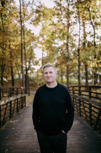 Toby Goostree, a white man with blond hair, stands with his hands in his pockets. He's wearing all black and standing on a wooden bridge with autumn trees behind him.