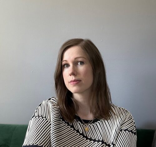 Brooke Middlebrook, a white woman with brown hair. She's sitting on a green couch and wearing a black shirt with a looping white design. The wall behind her is a neutral color.