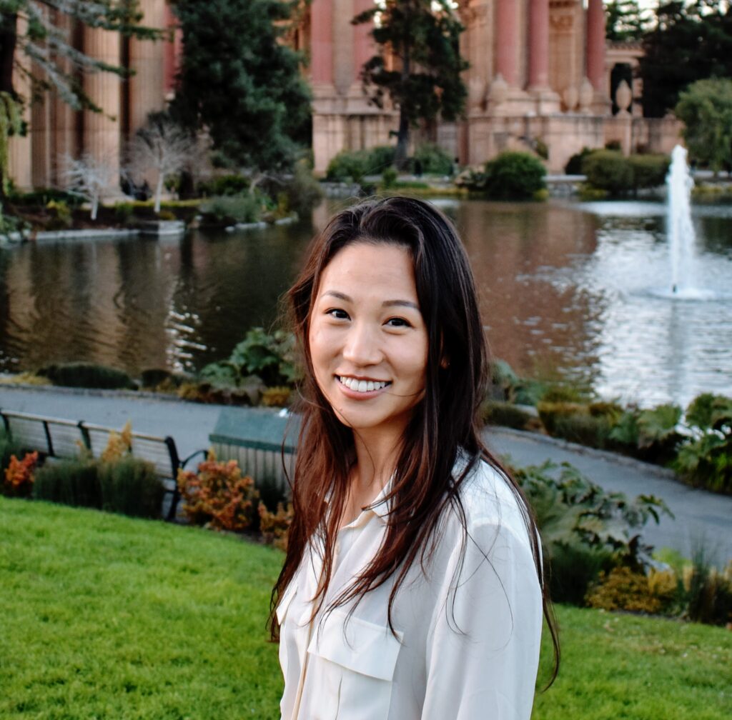 Catherine Niu, an Asian American woman with long highlighted hair, smiles in front of a pond and garden. She is wearing a white shirt.