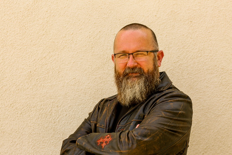 Jonathan Alexander, a white man with a buzz cut and dark and gray beard stands with arms folded in front of a cream-colored wall. He is wearing a leather jacket .