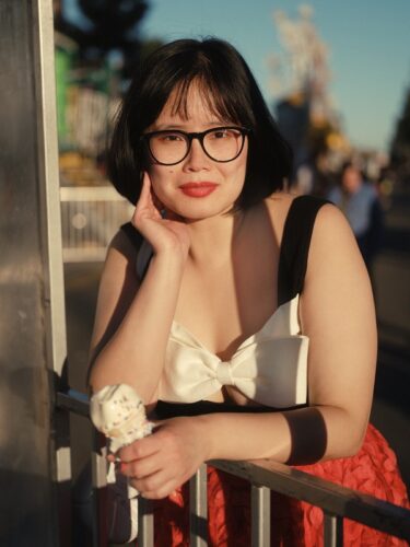 Dorothy Chan, an Asian nonbinary femme person, with dark short hair, dark glasses, and a dress with black shoulder straps, a white bow at the chest, and a red skirt. She is outside leaning on a railing with her hand near her cheek and holding an ice cream cone in the other.