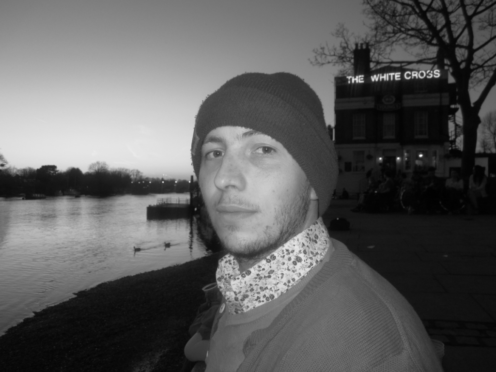 Daniel Fraser looks directly at the camera in a black-and-white photo. Behind him is a body of water and the silhouettes of a building and a tree.