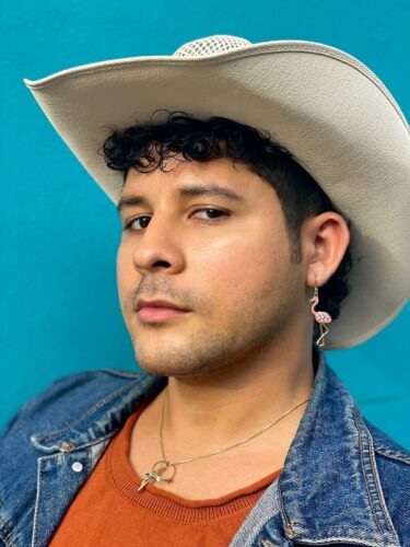 Edgar Gomez, a Latinx man with a cowboy hat, wears a jean jacket, an orange T-shirt, and a necklace with a charm. The backdrop is bright blue.