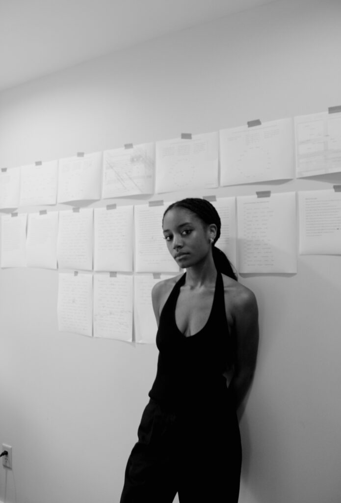 Kailah Figueroa, a Black woman with hair pulled back, leans against a wall covered in papers