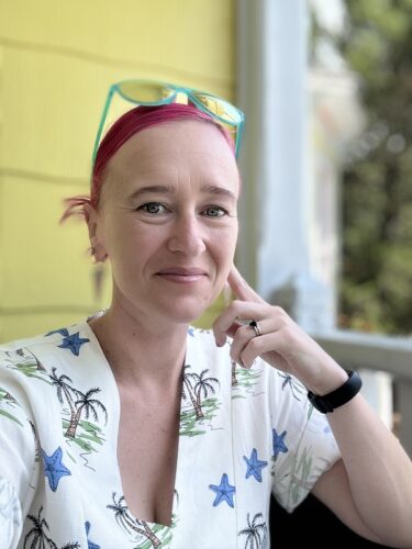 Gwen E. Kirby, a white woman wearing a shirt with palm trees and starfish. She has a pink headband and glasses with light-blue frames on her head and a black watch on one wrist. She is sitting on the porch of a building with yellow siding.