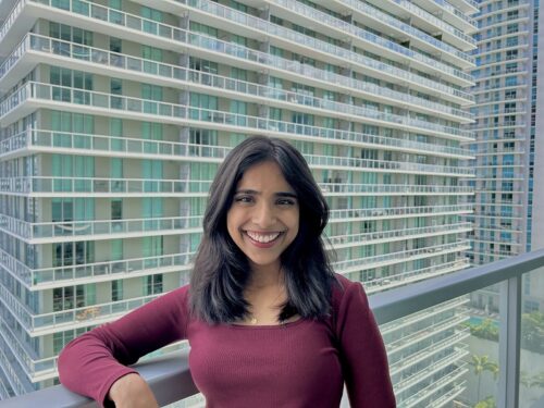 A Malayali American woman, Carolene Kurien stands on a balcony with a tall balconied building behind her. She is smiling and wearing a burgundy long-sleeve shirt.