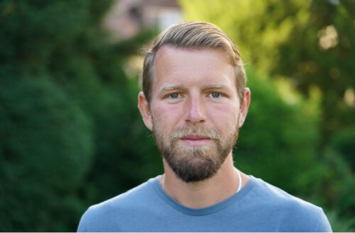 Vedran Husić, a white man wearing a light blue T-shirt. He has blondish hair and a beard and is standing in front of blurred-out trees.