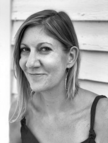 A black-and-white picture of Rebecca Bernard, a white woman with light-colored hair, smiling. She has on a tank top and long earrings, and she's standing in front of a house's siding.