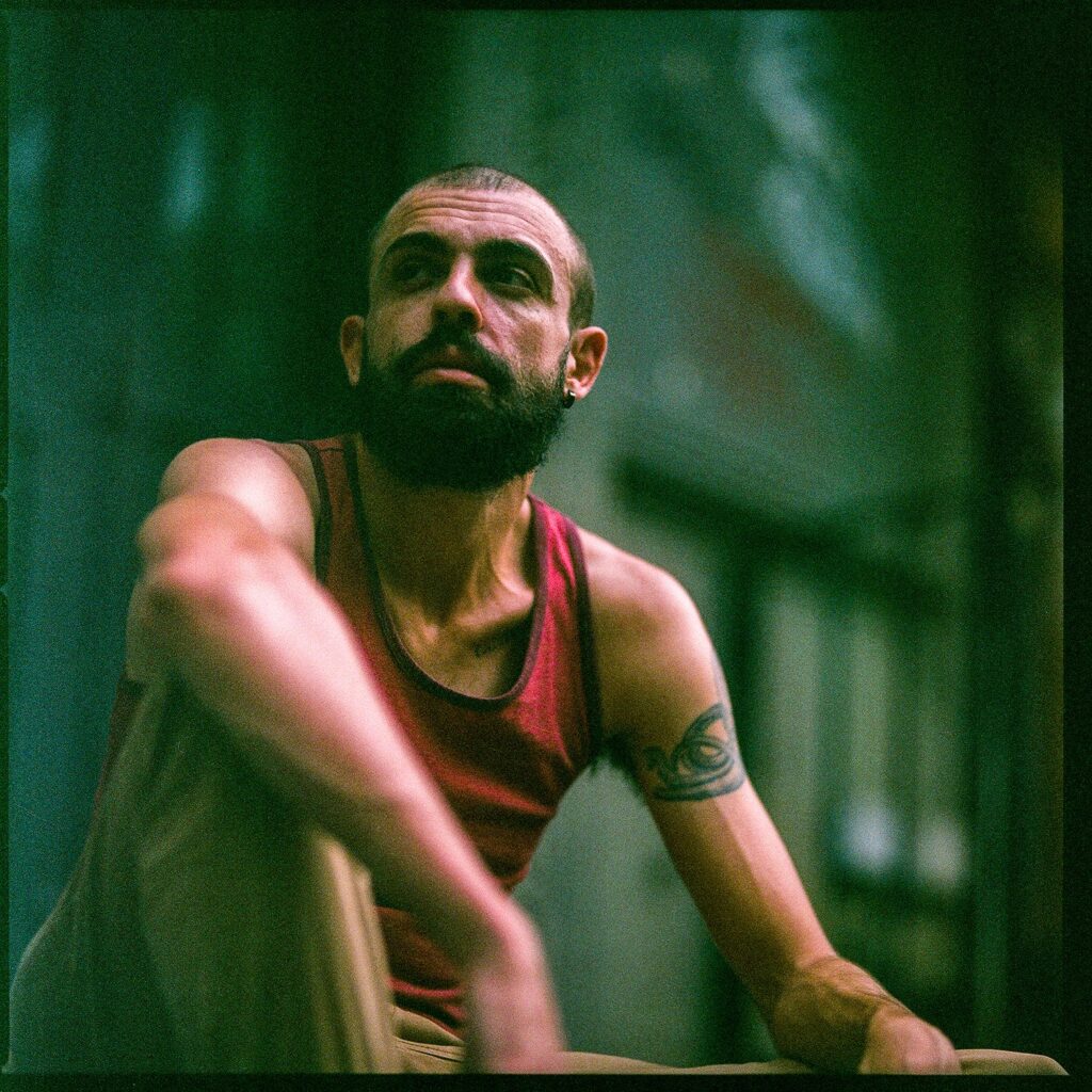 Alex Wells Shapiro sits in front of a blurry background with one knee up. He is wearing a red tank top.