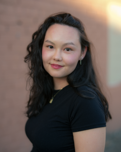 A woman from Hawai'i with dark hair and eyes, lipstick, and a black short-sleeved shirt; there's a blurred background behind her.